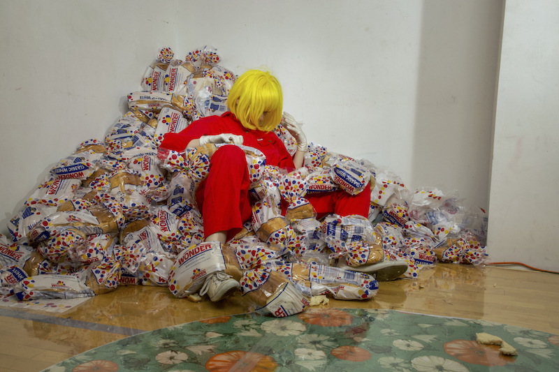 A performer, in a bright yellow wig and red jumpsuit, sits in a pile of Wonder Bread.